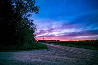 Road by trees against sky during sunset