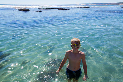 Portrait of shirtless boy wearing swimming googles standing in sea