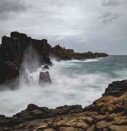 Scenic view of waves crashing at rocky coastline against sky