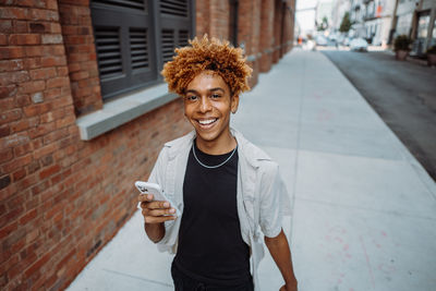 A happy young man in outerwear and eyewear walking sidewalk, holding cell phone