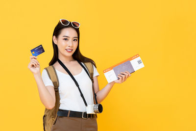 Young woman holding camera while standing against yellow background