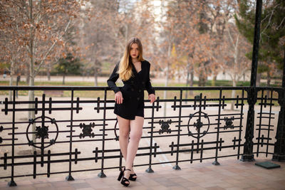 Portrait of confident beautiful young woman standing by railing against trees in park