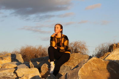 Portrait of young woman sitting on rock against sky