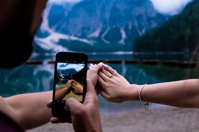 Midsection of man photographing with mobile phone while holding woman hand by lake