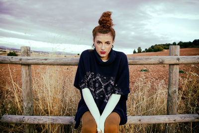 Portrait of young woman sitting on railing at field against sky