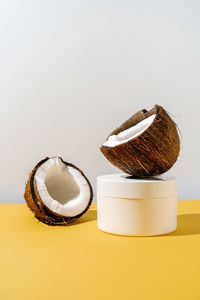 Two halves of coconut and a jar of cosmetics on a yellow and gray background.