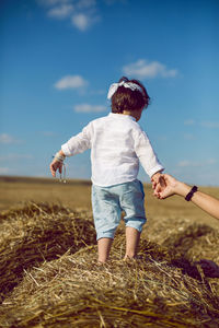 Boy child in a bandana and a white shirt stands back on a bale of hay and his father holds his hand