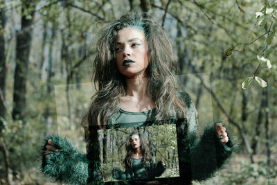 Serious young woman with spooky make-up holding picture frame while standing in forest