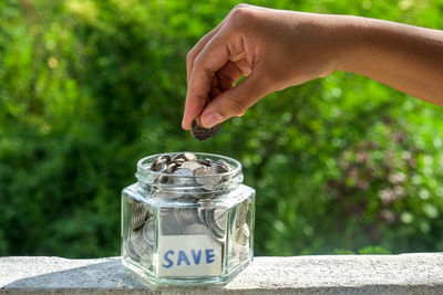 Close-up of human hand holding coins over jar with save label on retaining wall