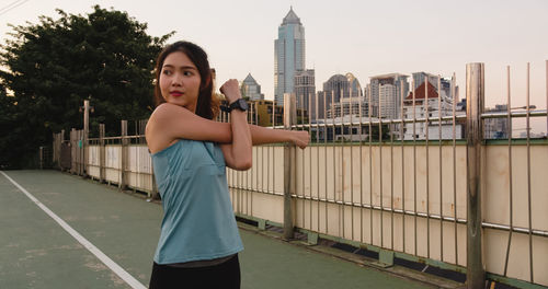 Beautiful young woman exercising while standing in city against sky