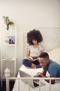 Smiling man using laptop computer while woman reading book on bed at home