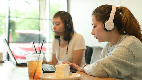 Two young women wearing headphones working with laptops in a cafe, cake and juice on the table.