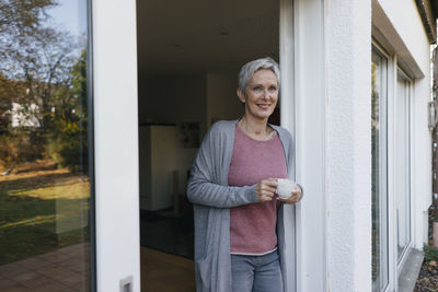 Smiling mature woman with cup of coffee at terrace door