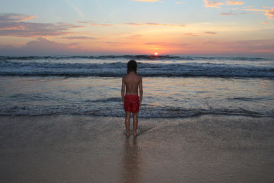 Rear view of boy standing at beach against sky during sunset