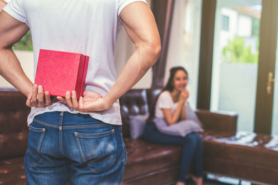 Midsection of boyfriend hiding gift while girlfriend sitting on sofa at home