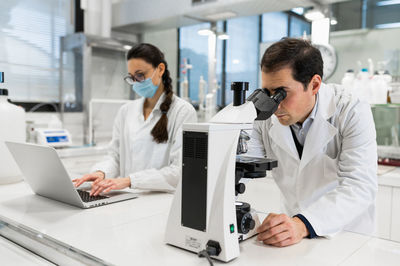 Concentrated male scientist using microscope and analyzing chemical samples while female colleague working on laptop in modern laboratory