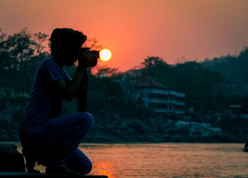 Side view of man sitting by lake against orange sky