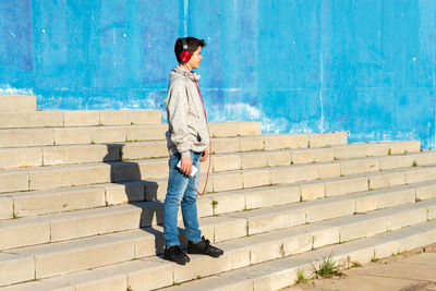Teen listening music by headphones while standing against blue wall