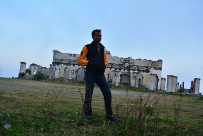 Man with hands in pocket standing on field against old ruin and blue sky