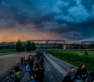 People sitting on bridge over river against cloudy sky