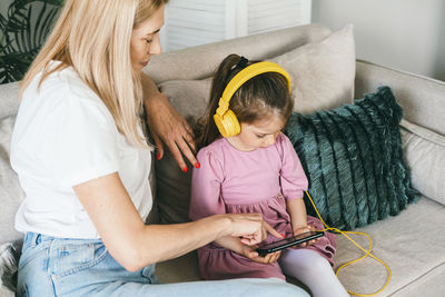 Little girl in yellow headphones sits on a sofa with a phone.