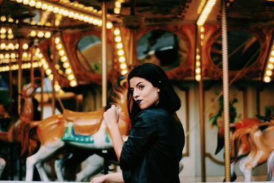 Portrait of young woman looking at illuminated carousel in amusement park