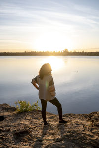 Rear view of young woman standing at beach against sky during sunset