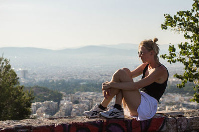 Young woman sitting on retaining wall against clear sky