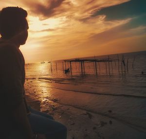 Young man sitting at beach against sky during sunset