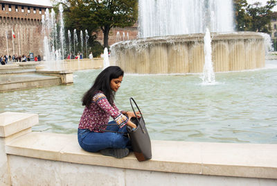 Full length of woman holding purse sitting by fountain