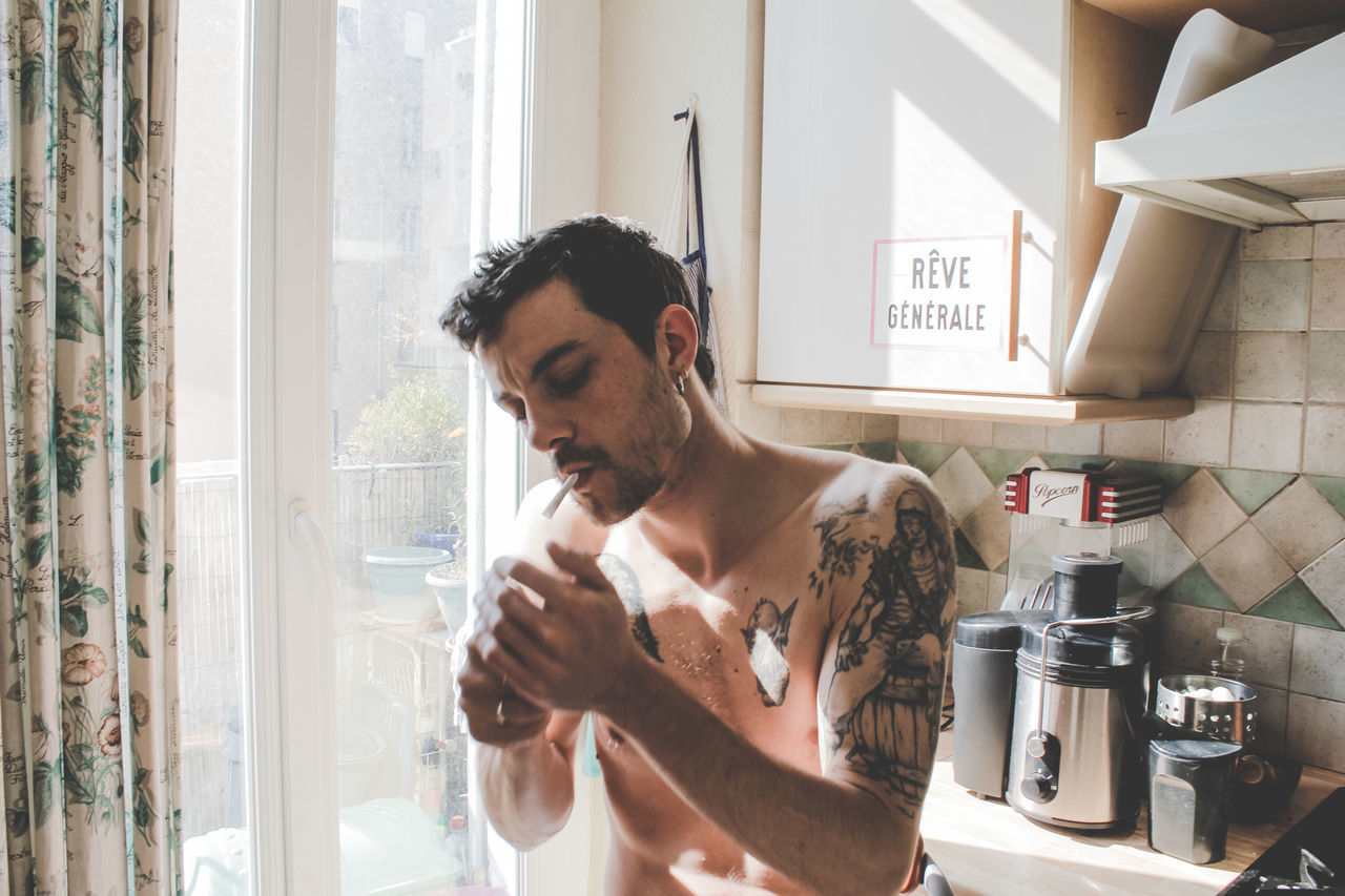 Shirtless man smoking cigarette by window