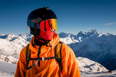 Portrait of a young man in a ski mask, stands in a ski resort against the backdrop of mountains and