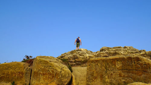 Low angle view of man standing on rock against clear blue sky