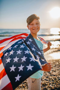 Portrait of boy holding american flag while standing at beach during sunset
