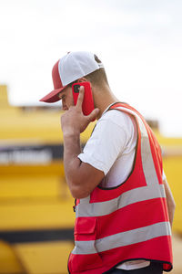 Side view of male worker in uniform having phone conversation while standing in agricultural field with combine harvester in countryside on summer day