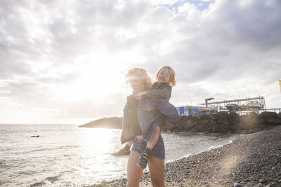 Mother piggybacking cute son while walking at beach against cloudy sky during sunset