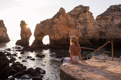 Anonymous female in hat sitting on stone and admiring sea and cliffs at sundown in ponta da piedade in algarve, portugal
