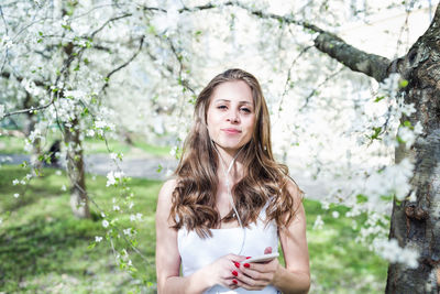 Young woman smiling and listening to music or podcast via earphones under blooming trees. spring par