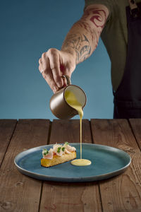 Hand pouring sauce on blue plate with rutabaga and lardo on wooden table with blue background