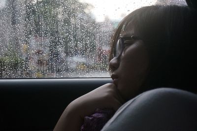 Side view of thoughtful young woman by wet window glass in car