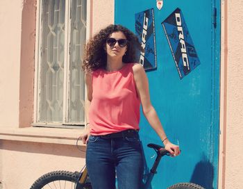 Portrait of young woman wearing sunglasses standing against house during sunny day