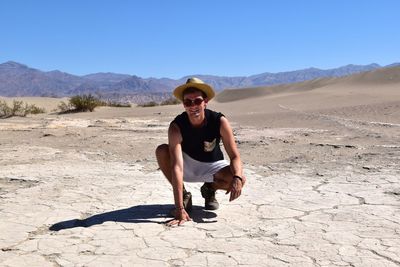 Portrait of young man wearing sunglasses crouching on arid landscape against clear blue sky