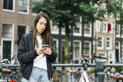 Side view of concentrated ethnic female traveler with long dark hair in casual clothes messaging on mobile phone while standing near parked bicycle on bridge during trip in amsterdam