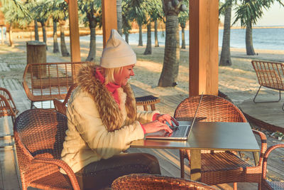 A woman in a sweater and a fur coat is sitting on the terrace of a cafe with a laptop.