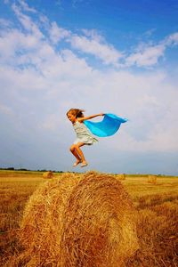 Girl jumping over hay bale on field against sky