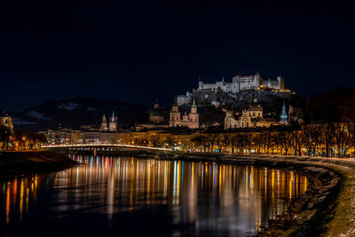 Illuminated buildings by river against sky at night