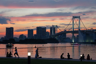 Silhouette of suspension bridge over river against cloudy sky