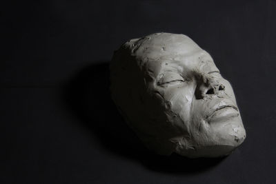 Clay sculpture, with closed eyes. face whit shadows, liyng on black floor.