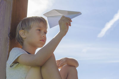 Boy holding paper airplane against cloudy sky