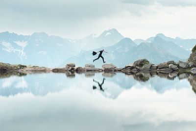 Man reflecting on calm lake while jumping mountains against cloudy sky